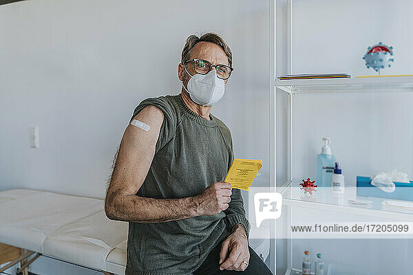 Männlicher Patient mit Schutzmaske und Verband am Arm  der den Impfpass hält  während er im Untersuchungsraum sitzt