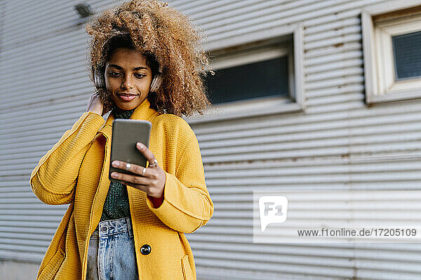 Lächelnde Afro-Frau mit Hand im Haar  die Musik hört  während sie ein Mobiltelefon an der Wand benutzt