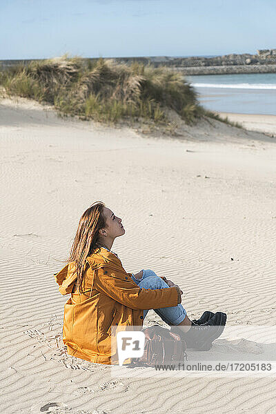 Junge Frau umarmt Knie beim Sitzen am Strand während eines sonnigen Tages