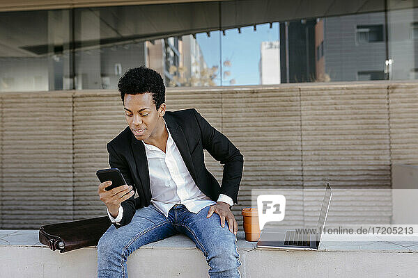 Mittlerer erwachsener Geschäftsmann  der sein Smartphone benutzt  während er auf einer Stützmauer sitzt