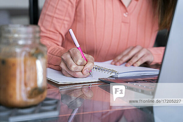 Junge Frau schreibt in Tagebuch  während sie am Esstisch im Heimbüro sitzt