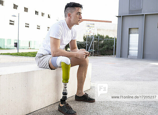 Behinderter Mann schaut weg  während er mit seinem Smartphone auf einer Stützmauer sitzt