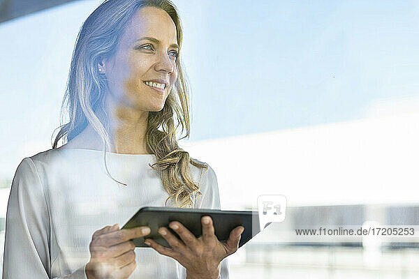 Lächelnde Frau mit digitalem Tablet  die wegschaut  während sie am Fenster im Büro steht