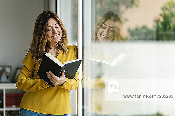 Lächelnde Frau  die ein Buch liest und sich zu Hause an ein Glasfenster lehnt