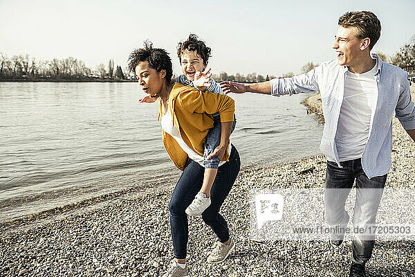 Mother piggybacking cheerful boy while running by lakeshore
