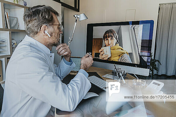 Älterer Arzt im Gespräch mit einem Patienten per Videoanruf über einen Computer  während er in der Arztpraxis sitzt