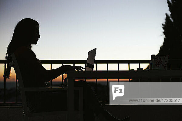 Silhouette einer Frau  die bei Sonnenaufgang auf dem Balkon am Laptop arbeitet