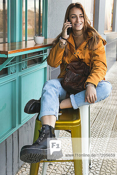Lächelnde Frau  die auf einem Hocker in einem Straßencafé sitzt und mit ihrem Handy telefoniert
