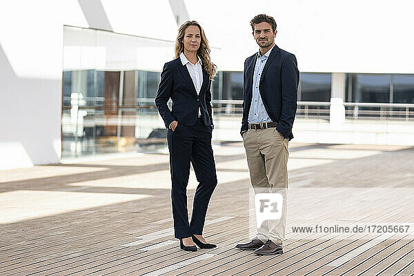 Confident business people standing with hands in pockets at office building terrace