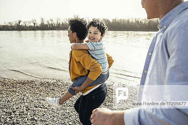 Fröhlicher Junge genießt es  von seiner Mutter huckepack genommen zu werden  während sein Vater am Seeufer spazieren geht