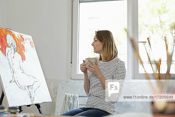 Frau mit Kaffeetasse und Blick auf ein Gemälde  während sie im Heimstudio sitzt