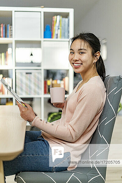 Junge Frau mit digitalem Tablet lächelnd und hält Kaffeetasse sitzen zu Hause