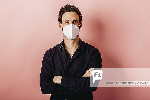 Mittlerer erwachsener Geschäftsmann mit FFP2-Gesichtsmaske  stehend mit verschränkten Armen vor farbigem Hintergrund