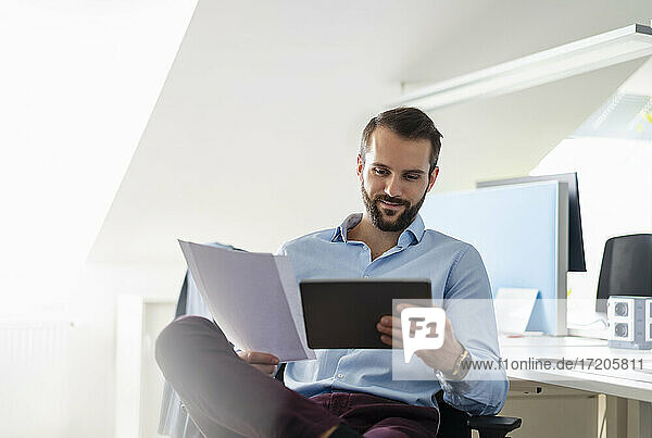 Lächelnder Unternehmer mit digitalem Tablet und Papier im Büro sitzend