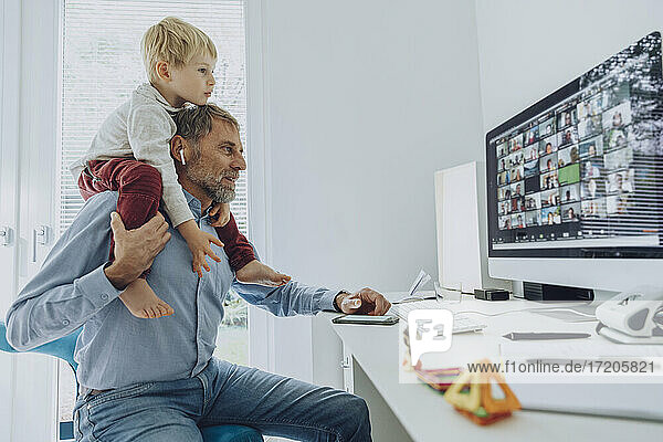 Vater trägt seinen Sohn auf der Schulter  während er an einer Videokonferenz im Büro teilnimmt