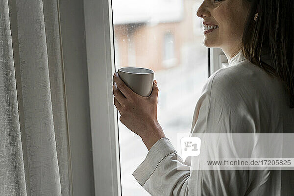 Lächelnde Frau mit Kaffee am Fenster stehend