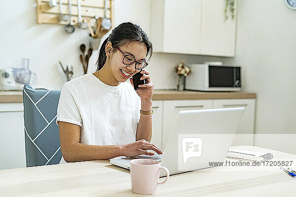 Junge Frau spricht mit dem Handy  während sie einen Laptop in der Küche benutzt