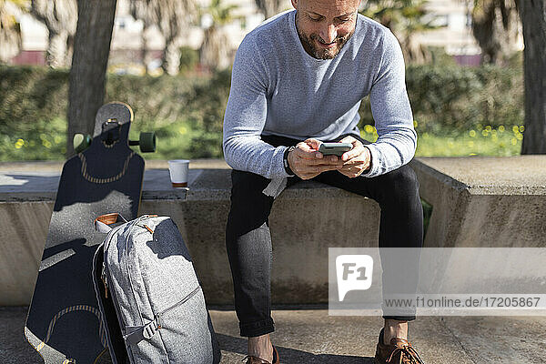 Männlicher Berufstätiger  der sein Smartphone benutzt  während er auf einer Betonbank im Park sitzt