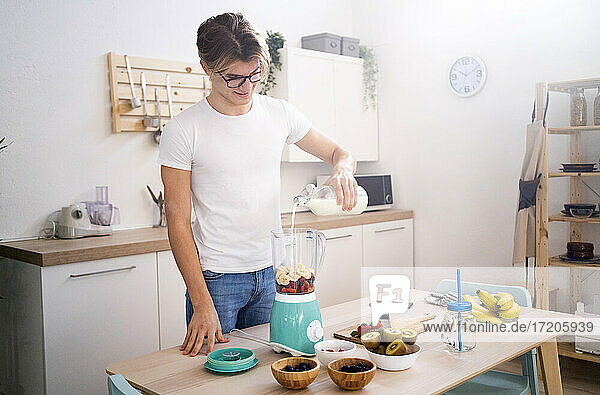 Junger Mann gießt Milch in den Mixer  während er in der Küche einen Smoothie zubereitet