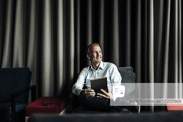 Lächelnder Geschäftsmann mit übereinander geschlagenen Beinen  der ein digitales Tablet auf einem Sessel in einer Büro-Cafeteria hält