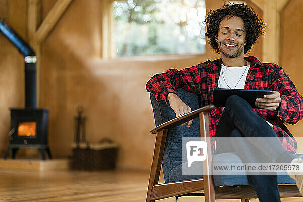 Lächelnder Mann  der ein digitales Tablet benutzt  während er in einem geräumigen Zimmer auf einem Sessel sitzt
