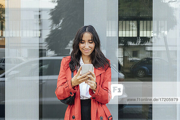 Smiling woman in red trench coat using smart phone while standing against glass wall