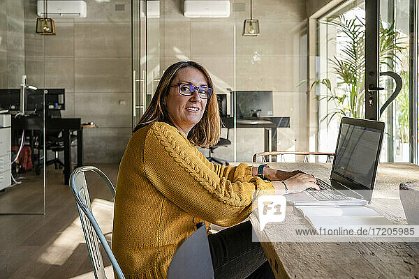 Mature female entrepreneur using laptop while sitting at conference table against glass door in office