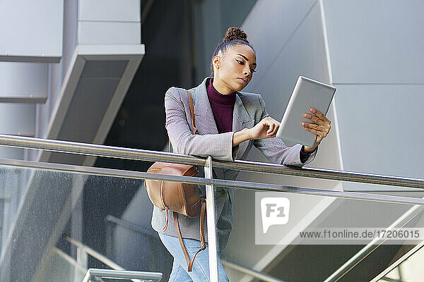 Eine Unternehmerin benutzt ein digitales Tablet  während sie sich an ein Geländer eines Bürogebäudes lehnt