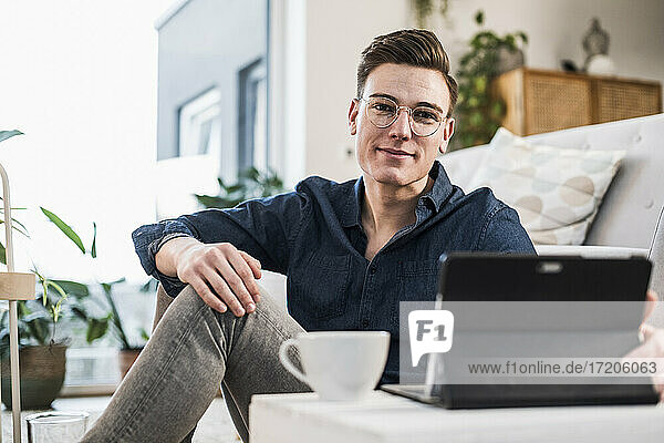 Lächelnder junger Mann mit Brille und Laptop im Wohnzimmer sitzend