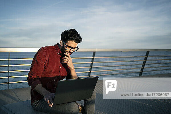 Nachdenklicher Geschäftsmann schaut auf seinen Laptop  während er auf einer Bank am Meer sitzt