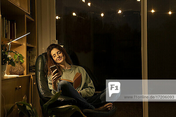 Smiling woman with book using smart phone while sitting at home