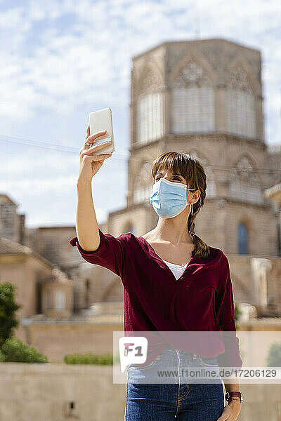 Frau  die während der Pandemie ein Selfie mit dem Handy vor einem Gebäude macht