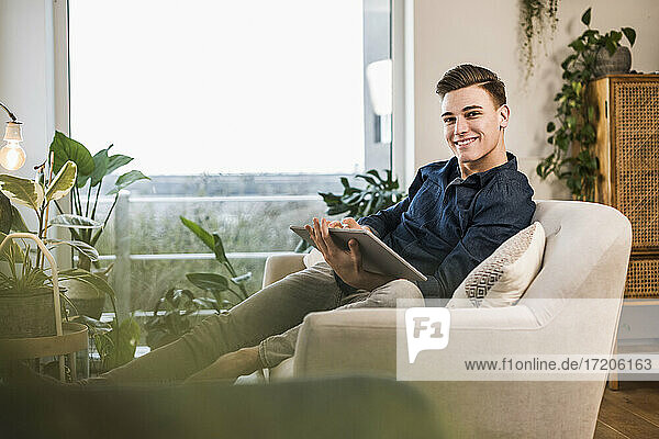 Lächelnder junger Mann mit digitalem Tablet auf dem Sofa im Wohnzimmer sitzend
