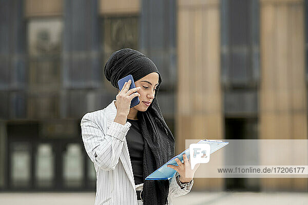 Young female entrepreneur talking on smart phone holding strategy while standing outdoors