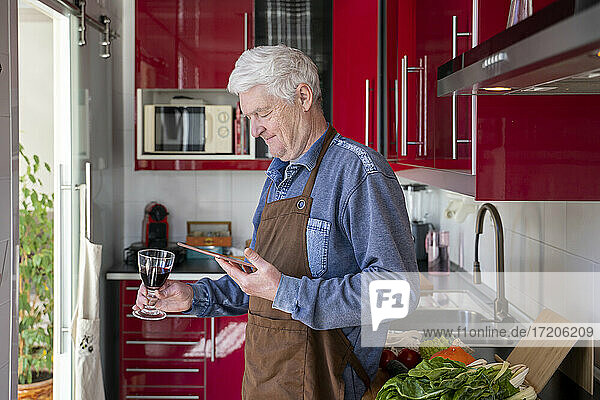 Älterer Mann hält ein Glas Wein in der Hand und benutzt ein digitales Tablet in der Küche