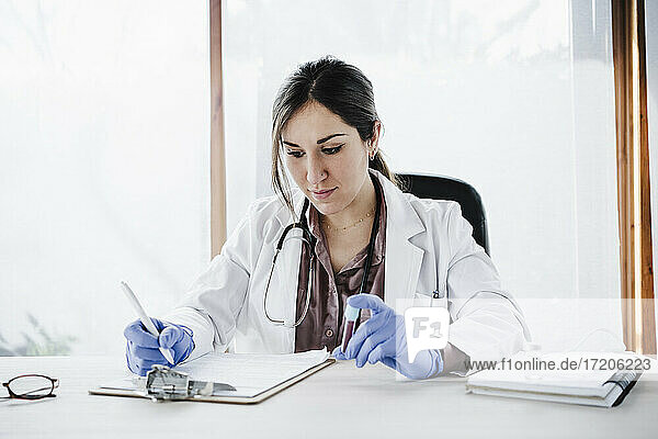 Weibliches medizinisches Fachpersonal schreibt Bluttestberichte  während es eine Blutprobe am Schreibtisch hält
