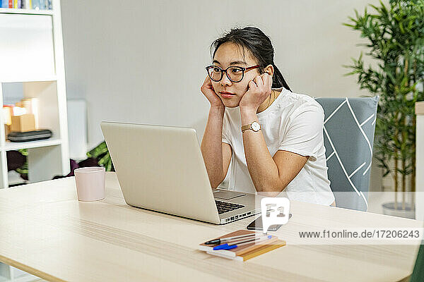 Junge nachdenkliche Frau sitzt mit Laptop auf dem Tisch zu Hause