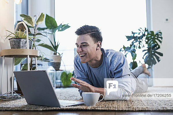 Lächelnder junger Mann  der auf einem Teppich liegt und bei einem Videoanruf über einen Laptop gestikuliert