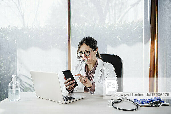 Lächelnde Ärztin  die ein Mobiltelefon benutzt  während sie am Schreibtisch im Krankenhaus sitzt