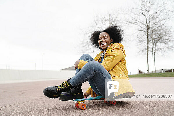 Lächelnde Frau auf dem Skateboard sitzend auf dem Fußweg gegen den klaren Himmel