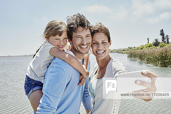 Lächelnde Frau nimmt Selfie mit Familie am See an einem sonnigen Tag