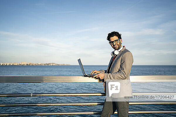 Smiling male business professional with laptop standing at railing by sea