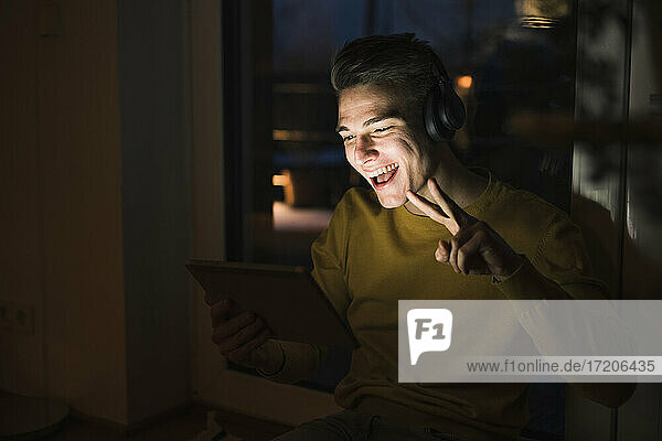 Fröhlicher Mann mit Kopfhörern  der bei einem Videoanruf das Friedenszeichen macht  während er im Wohnzimmer sitzt