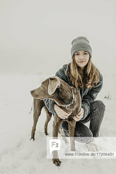 Porträt eines Mädchens im Teenageralter  das im Schnee hockt und einen Labrador Retriever umarmt