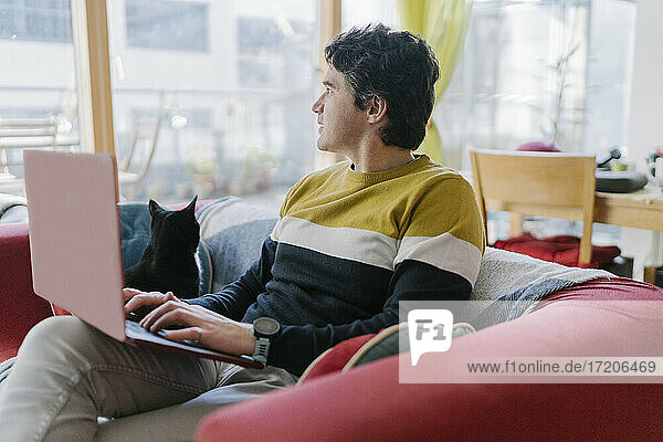 Mid adult man using laptop looking away while sitting on sofa at home
