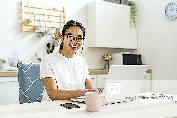 Lächelnde Frau sitzt am Tisch mit Laptop zu Hause