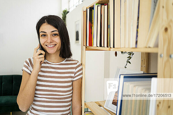 Lächelnde Frau  die am Bücherregal im Wohnzimmer telefoniert