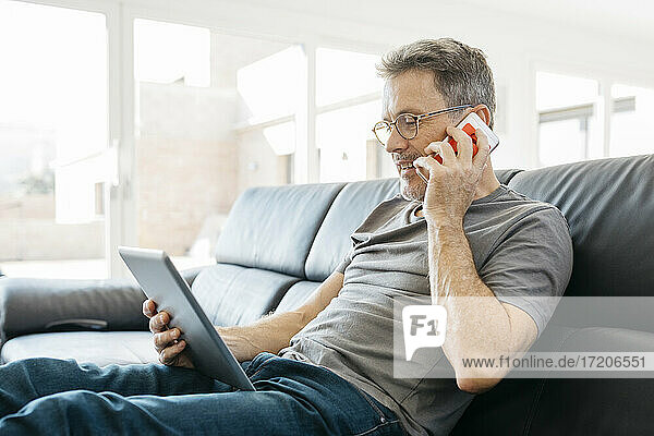 Mature man talking on mobile phone while using digital tablet on sofa in living room