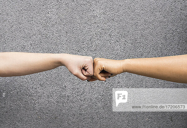 Arms of two young women fist bumping against gray background