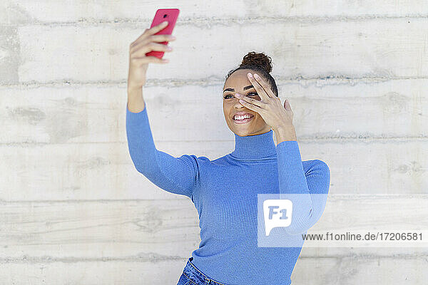 Lächelnde Frau gestikuliert  während sie ein Selfie durch ein an der Wand stehendes Mobiltelefon macht